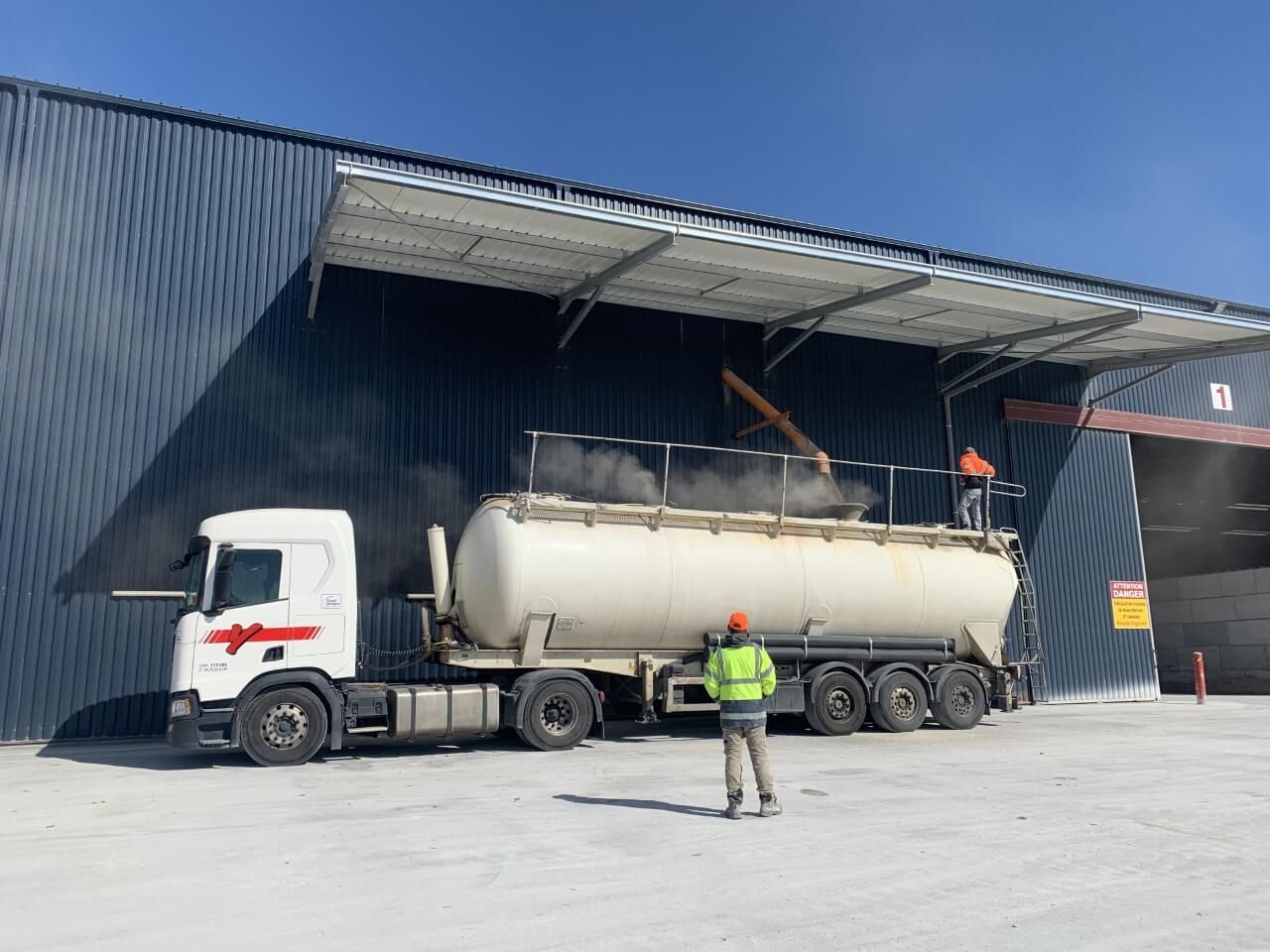 Chargement d'un conteneur cylindrique blanc dans un camion. Des ouvriers en tenue de sécurité sous un auvent bleu foncé.