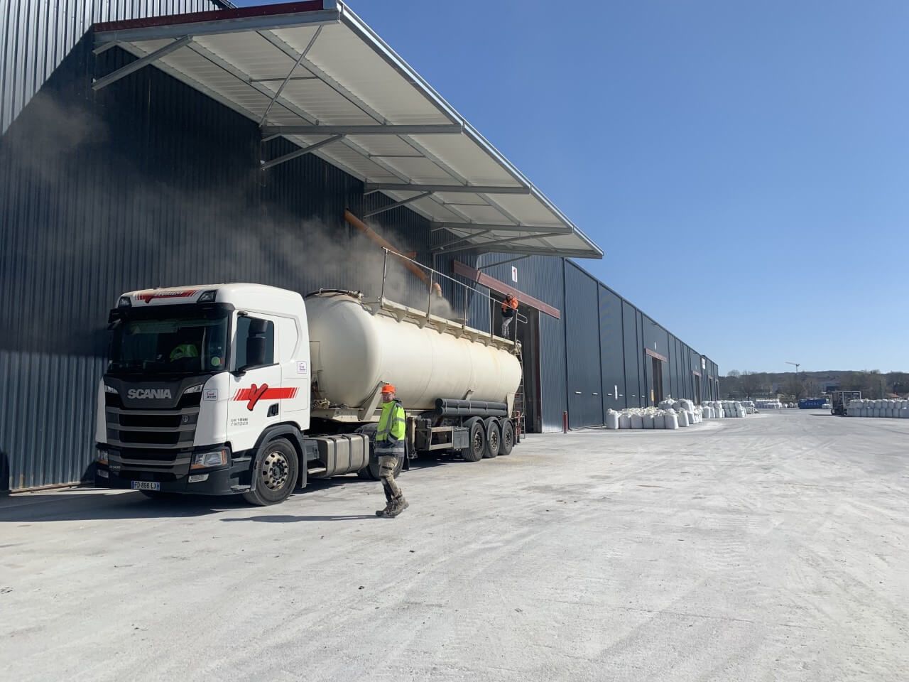 Un camion-citerne est chargé de poudre dans un entrepôt. Un ouvrier se tient à proximité, sous un ciel bleu.