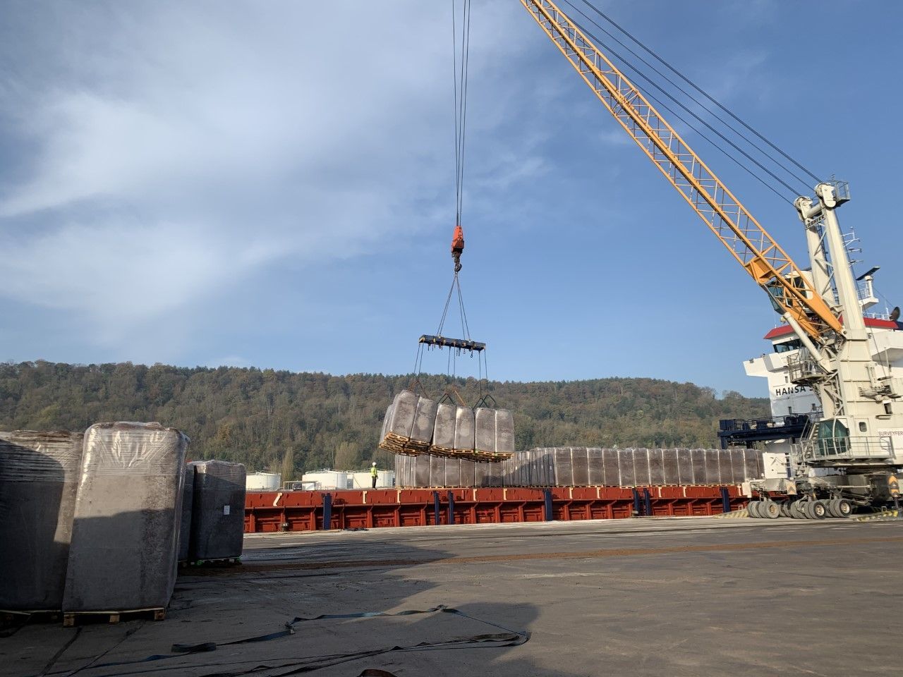 Grue chargeant une cargaison emballée sur un navire. Montagnes verdoyantes et ciel bleu en arrière-plan.