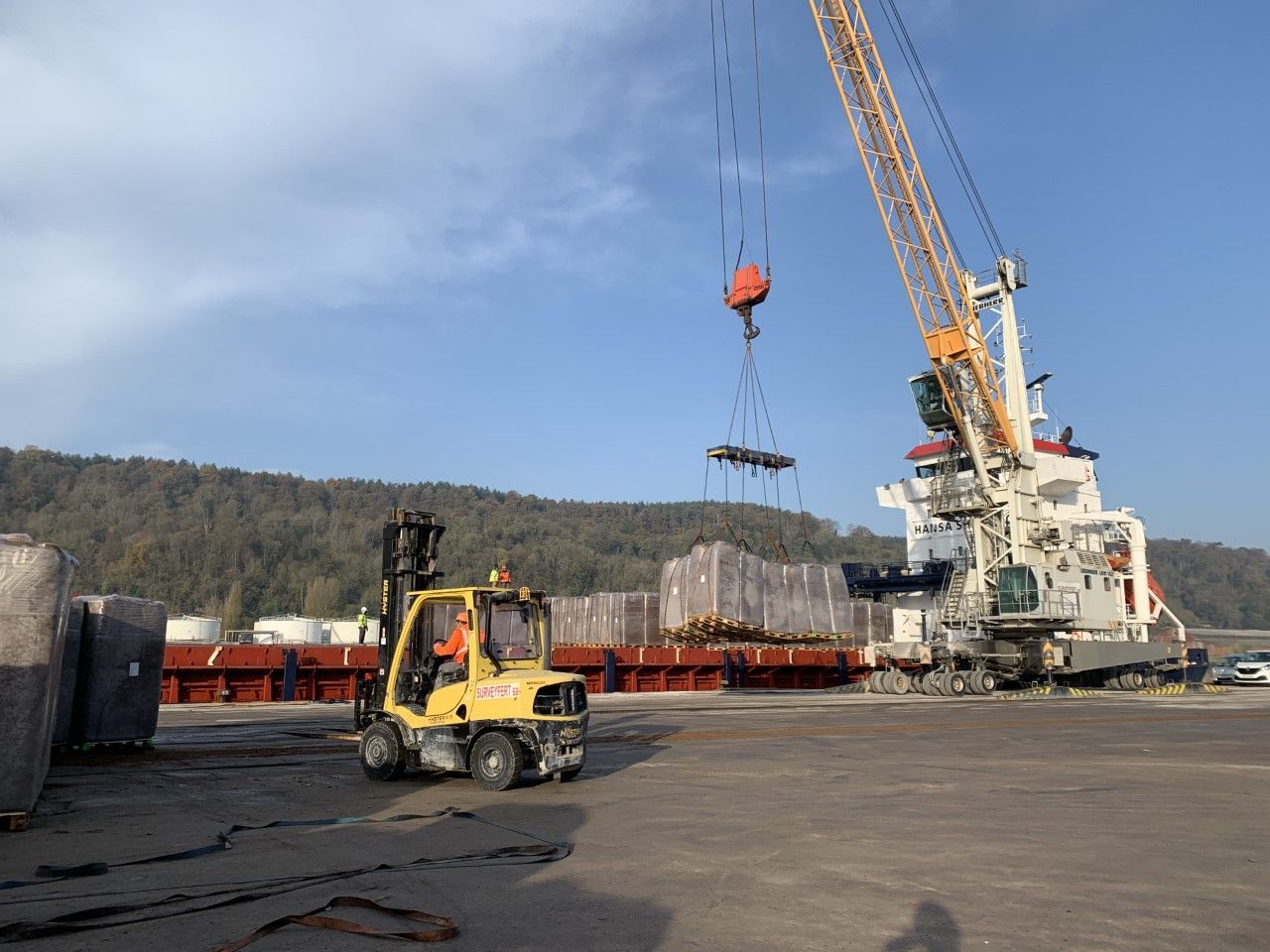 Un chariot élévateur et une grue déchargent la cargaison d'un navire dans un port. Ciel bleu avec forêt en arrière-plan.