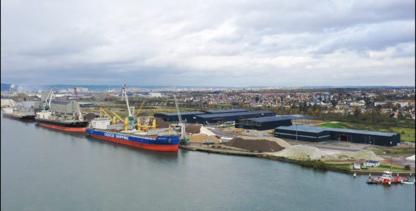 Navires amarrés dans un port, grues en cours de chargement, bâtiments en arrière-plan sous un ciel couvert.