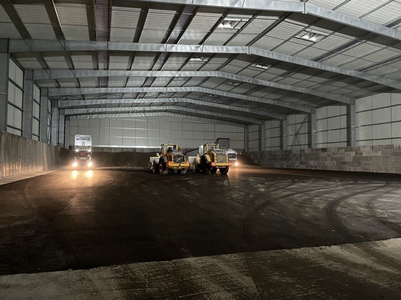 À l'intérieur d'un grand entrepôt : deux chargeurs et un camion, phares allumés, répandent du matériel sombre.