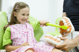 Una niña está sentada en una silla dental cepillándose los dientes con un cepillo de dientes de juguete.