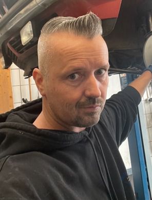 Eine Person in einem schwarzen Kapuzenpullover lehnt sich in ein Fahrzeug hinein; im Hintergrund ist ein blauer Aufzug oder Rahmen zu sehen.
