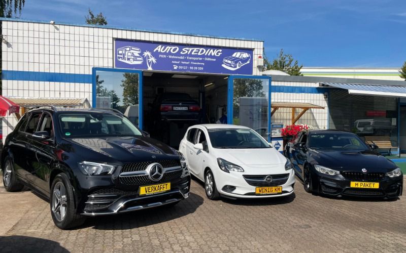 Drei Autos parkten vor einem blauen Autogeschäft, darunter ein schwarzer Mercedes und zwei kleine Schrägheckwagen.