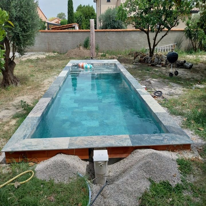 Piscine rectangulaire enterrée, entourée de dalles de pierre et d'une bordure en bois, dans un jardin.