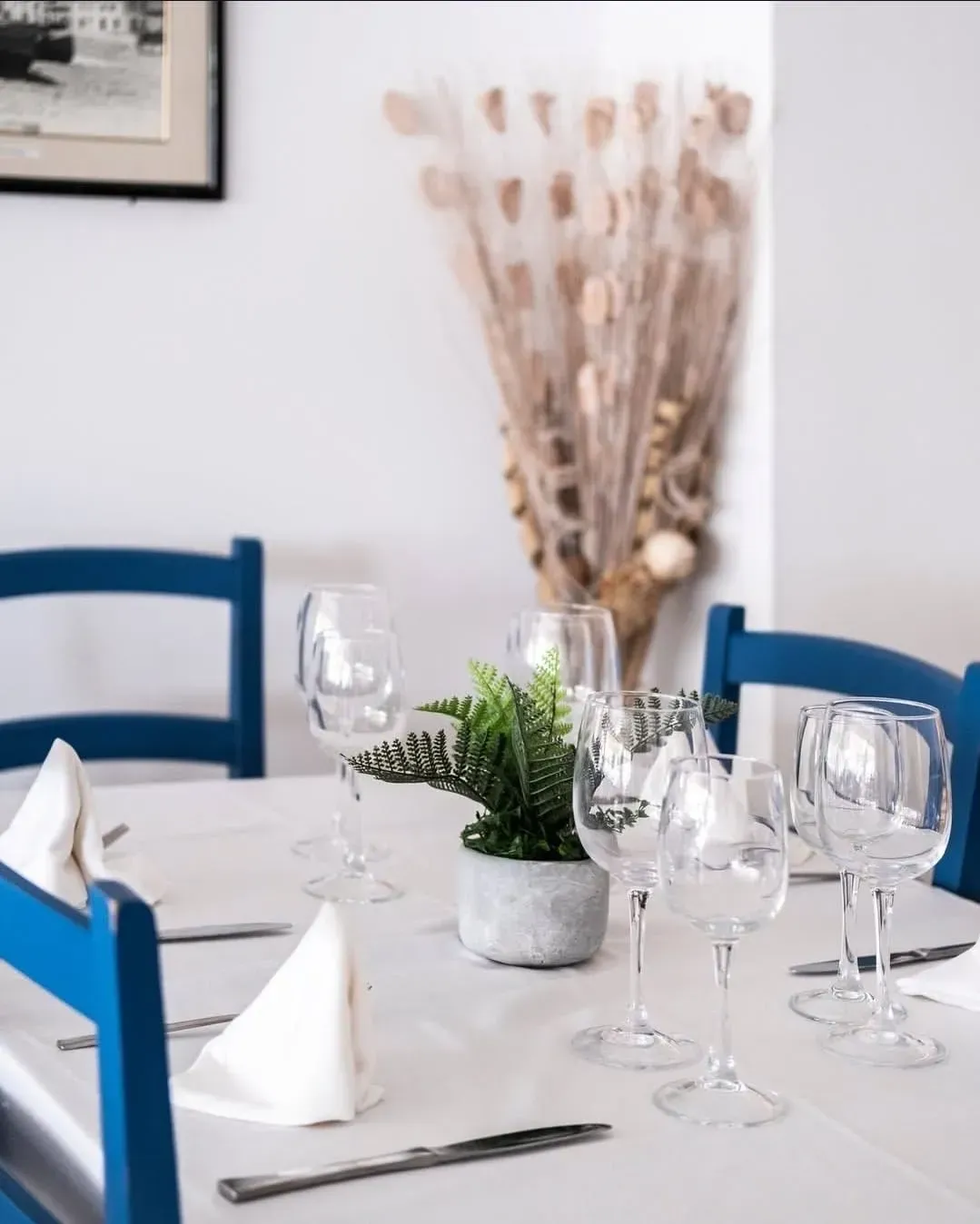 Una mesa de comedor con un mantel claro, sillas de madera azules, copas de vino, servilletas y una pequeña planta verde en maceta.