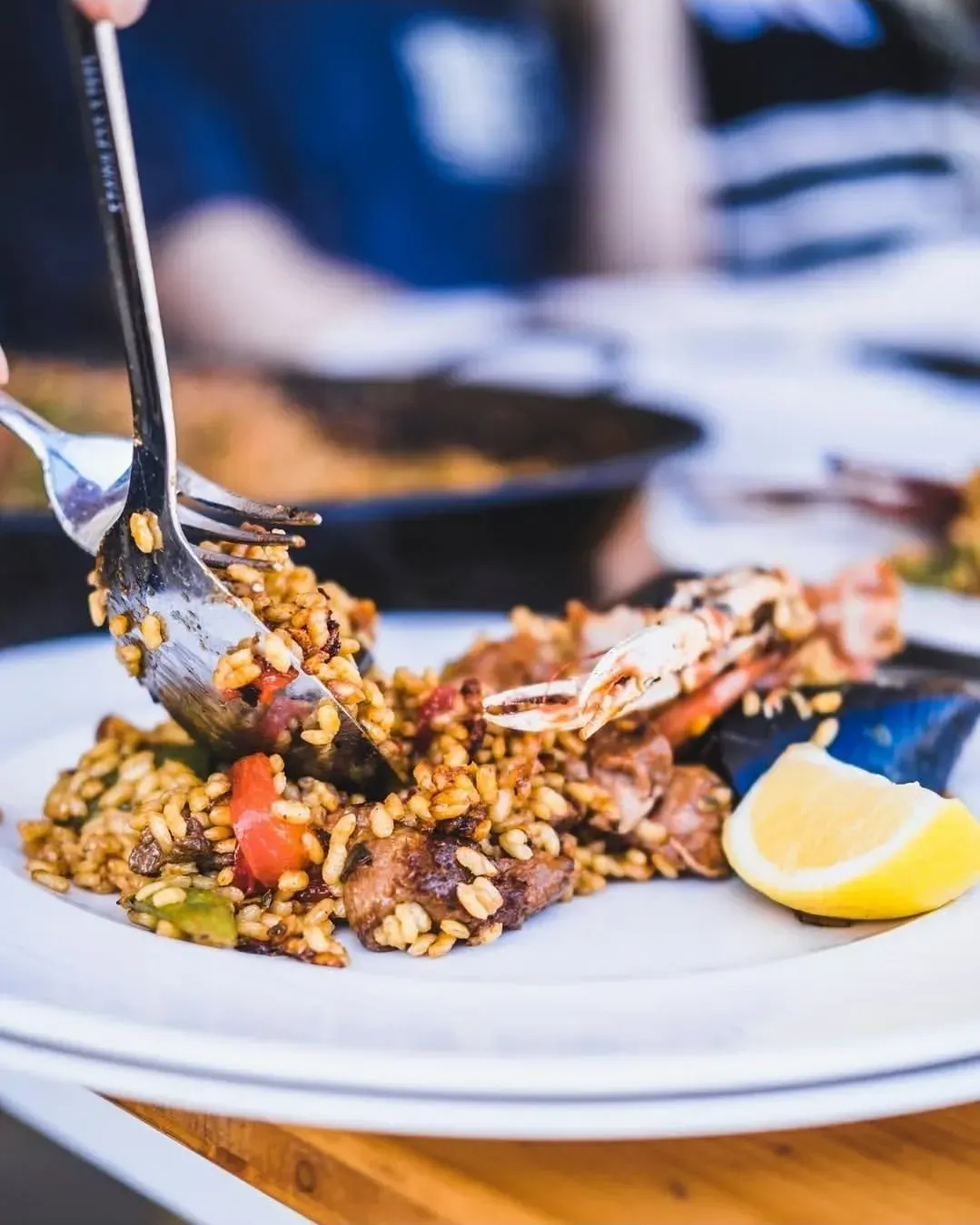 Una persona utiliza una cuchara y un tenedor para servir paella con marisco y una rodaja de limón en un plato blanco.