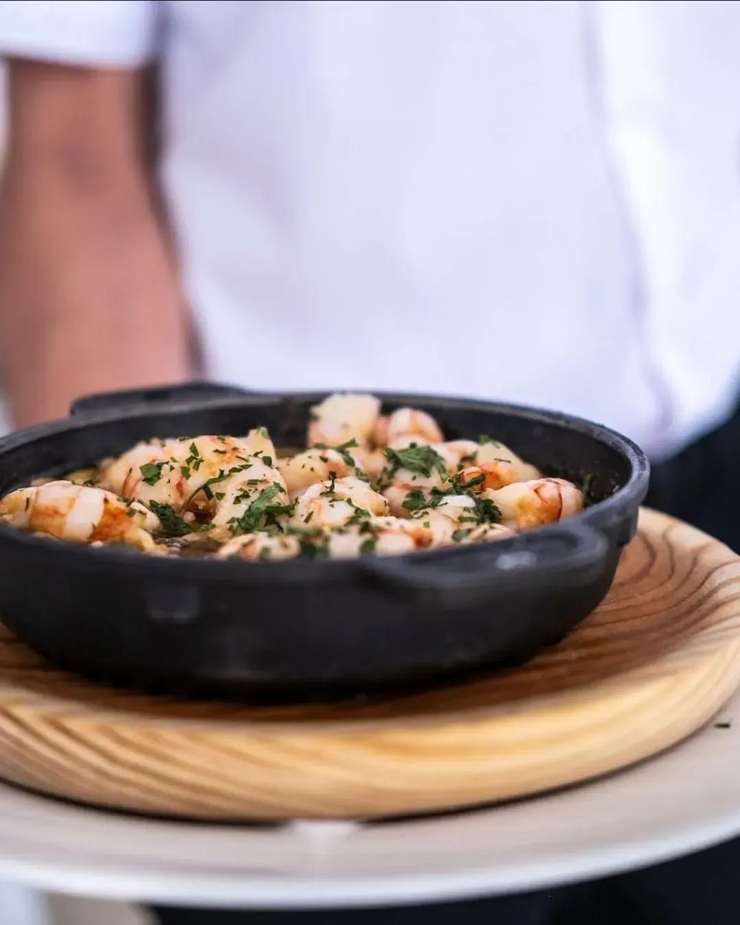 Un camarero sostiene en una bandeja de madera una sartén de hierro fundido negra llena de gambas cocidas y aderezadas con hierbas frescas.