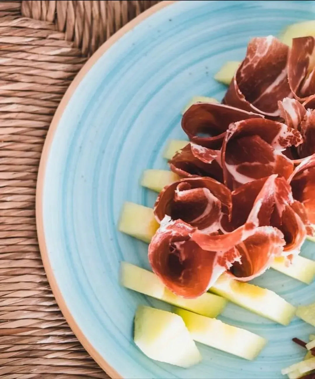 Prosciutto cortado en finas lonchas y doblado sobre trozos de melón cantalupo, servido en un plato azul estriado sobre una estera tejida.