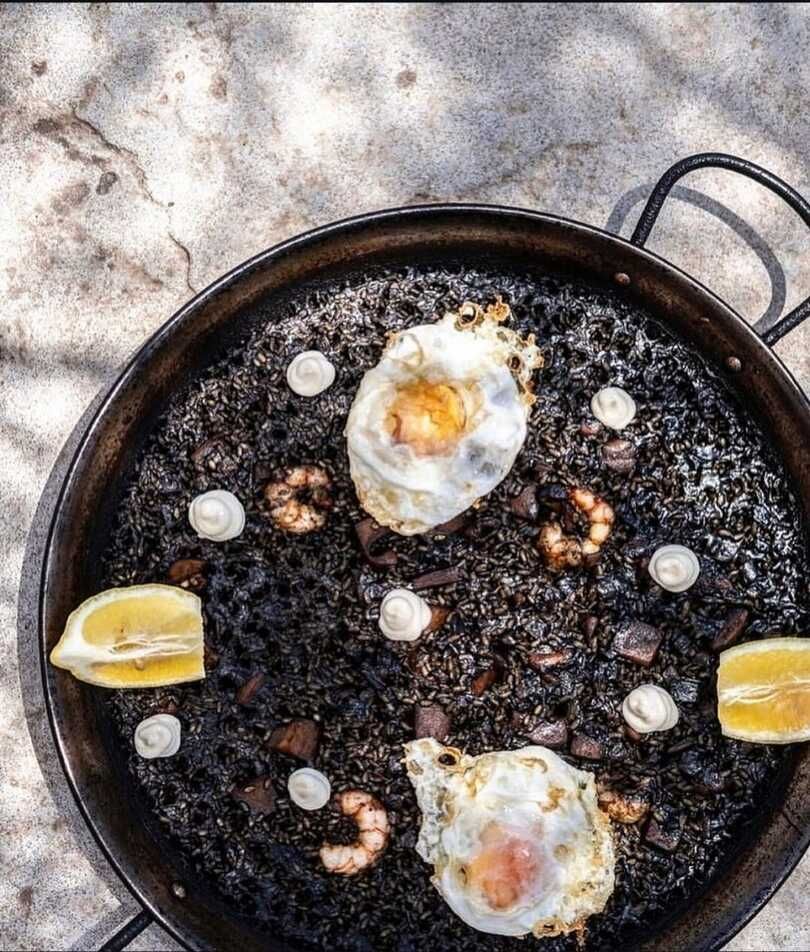 Una paellera rellena de arroz con tinta de calamar negra, cubierta con dos huevos fritos, gambas y rodajas de limón sobre una superficie de piedra.