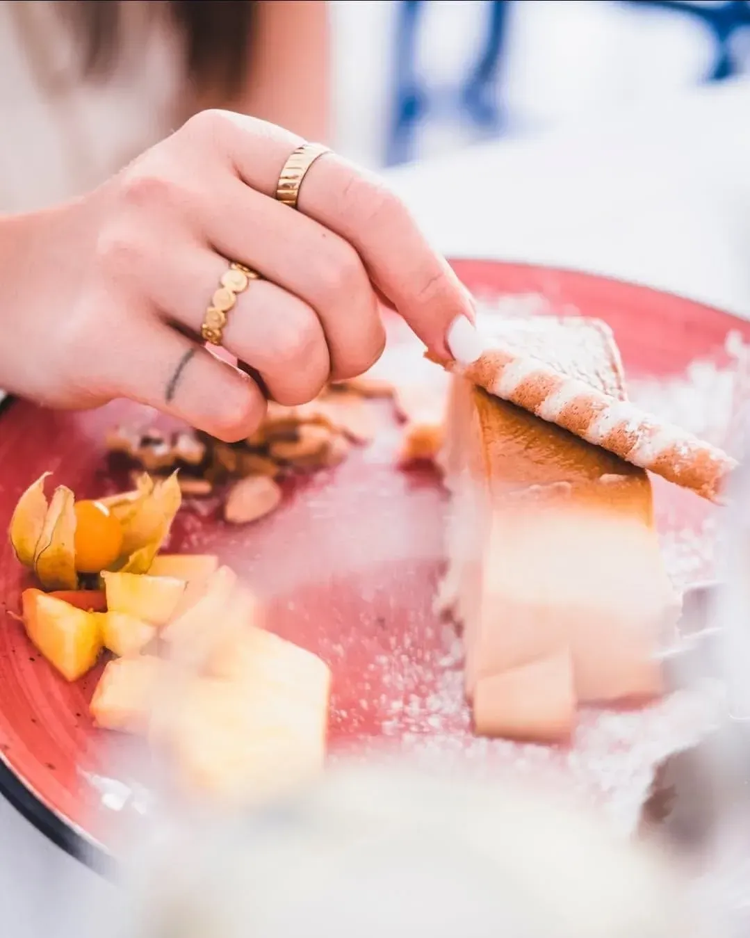 Una mano con anillos de oro decora un postre en un plato rojo con fruta en rodajas y una galleta.