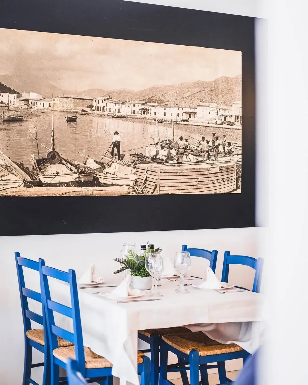 Una mesa para cuatro personas, puesta con manteles blancos y sillas azules, se encuentra debajo de una gran fotografía antigua enmarcada de un pueblo portuario.