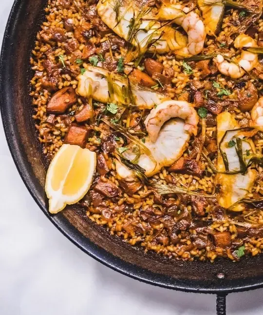 Una paella de marisco servida en una sartén de metal oscuro, adornada con hierbas aromáticas y una rodaja de limón fresco.
