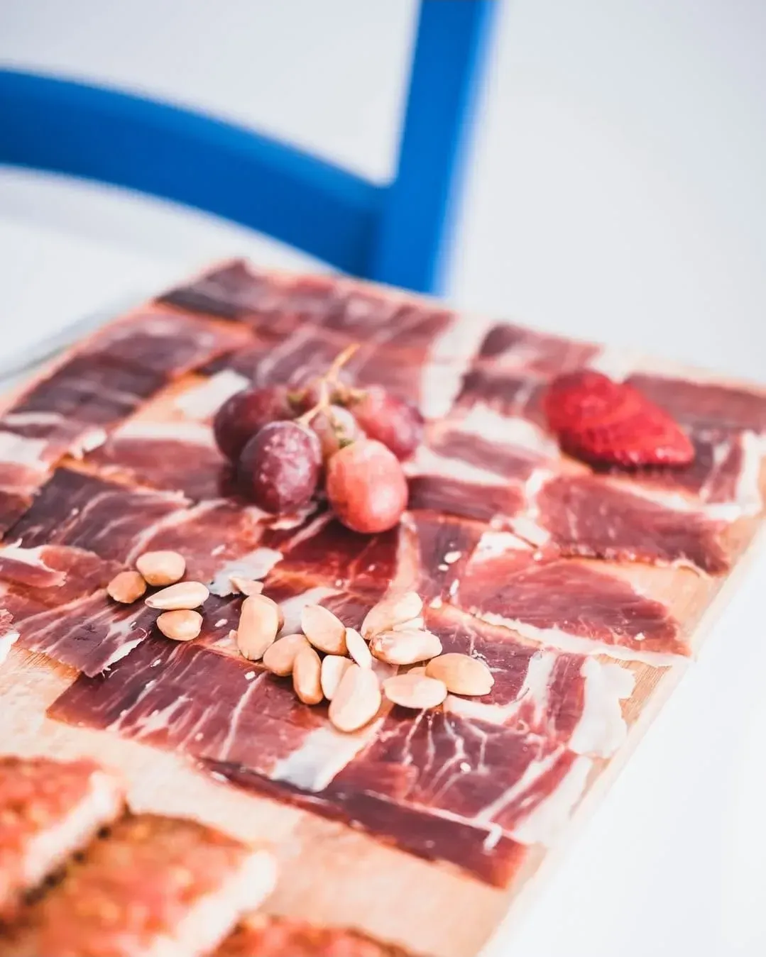Una tabla de madera para servir cubierta con lonchas de jamón curado, almendras, uvas y una fresa, con una silla azul al fondo.