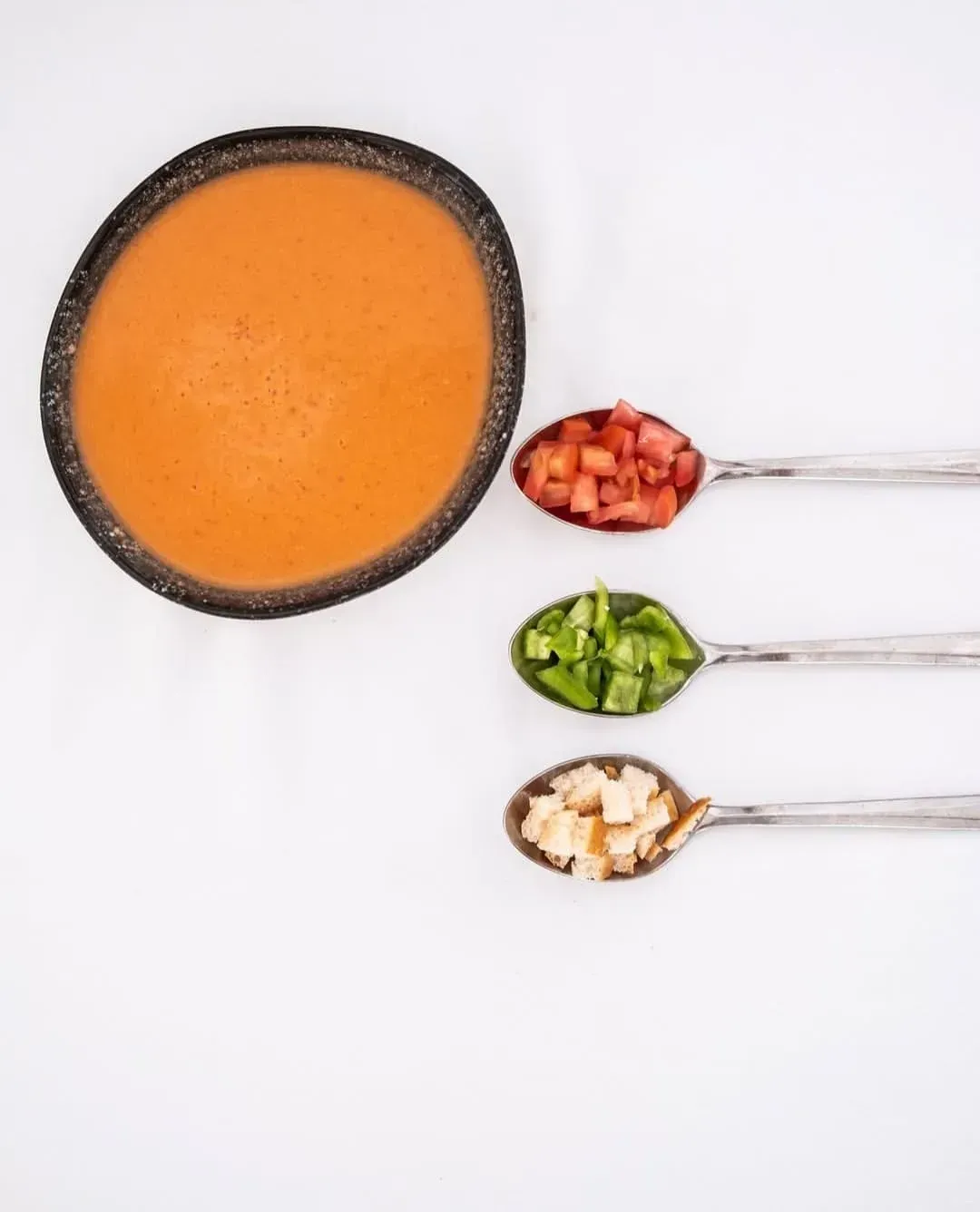 Un bol de gazpacho junto a tres cucharas llenas de tomate picado, pimiento verde y picatostes de pan sobre un fondo blanco.