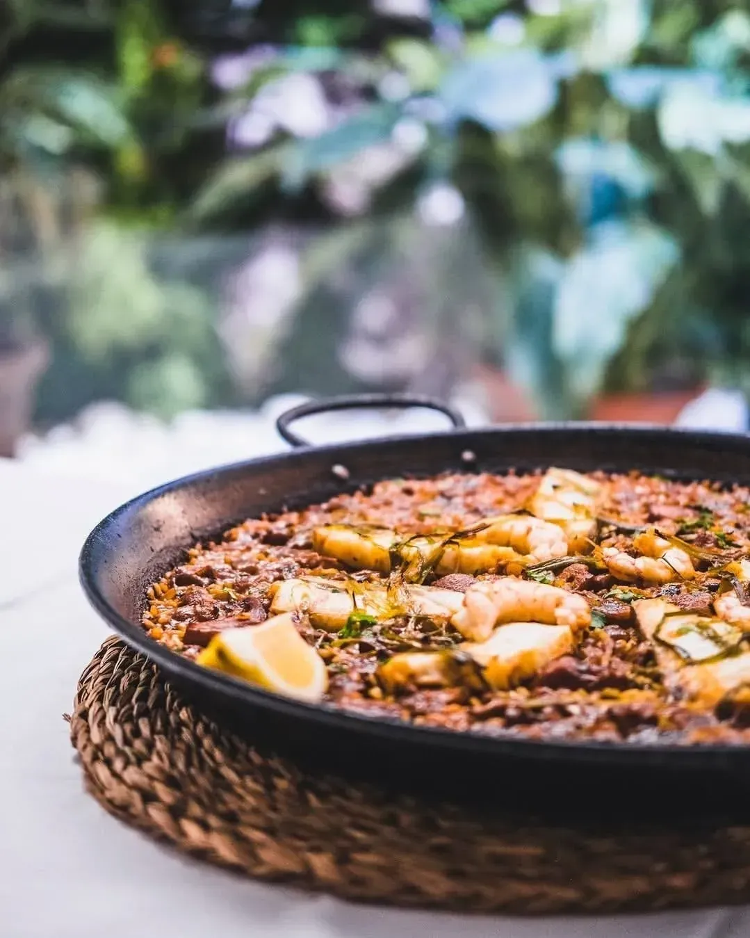 Una paella de marisco poco profunda con gambas y rodajas de limón, colocada sobre un mantel individual tejido al aire libre.