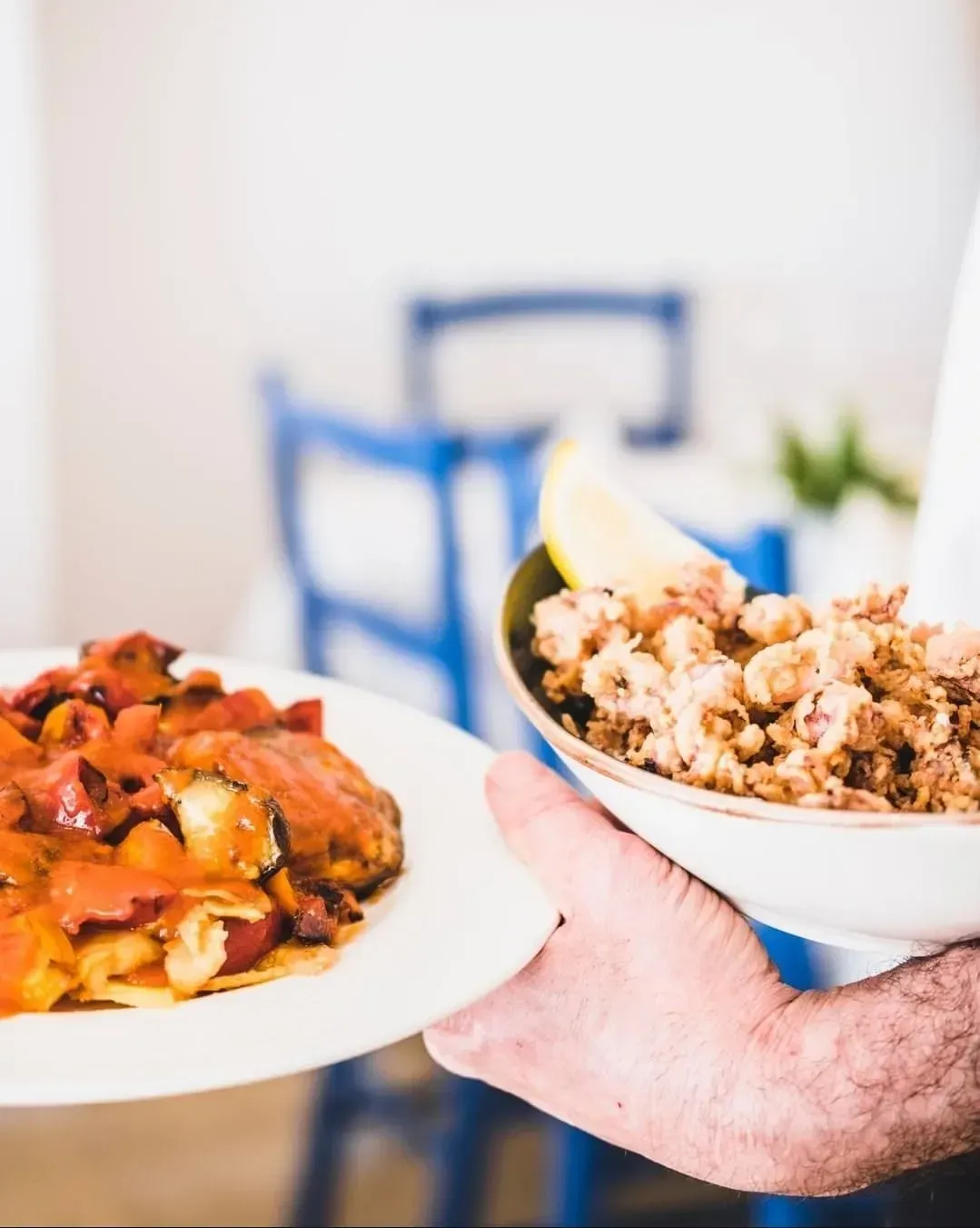 Una persona sostiene un plato de estofado y un tazón de calamares fritos con una rodaja de limón en un restaurante luminoso y espacioso.
