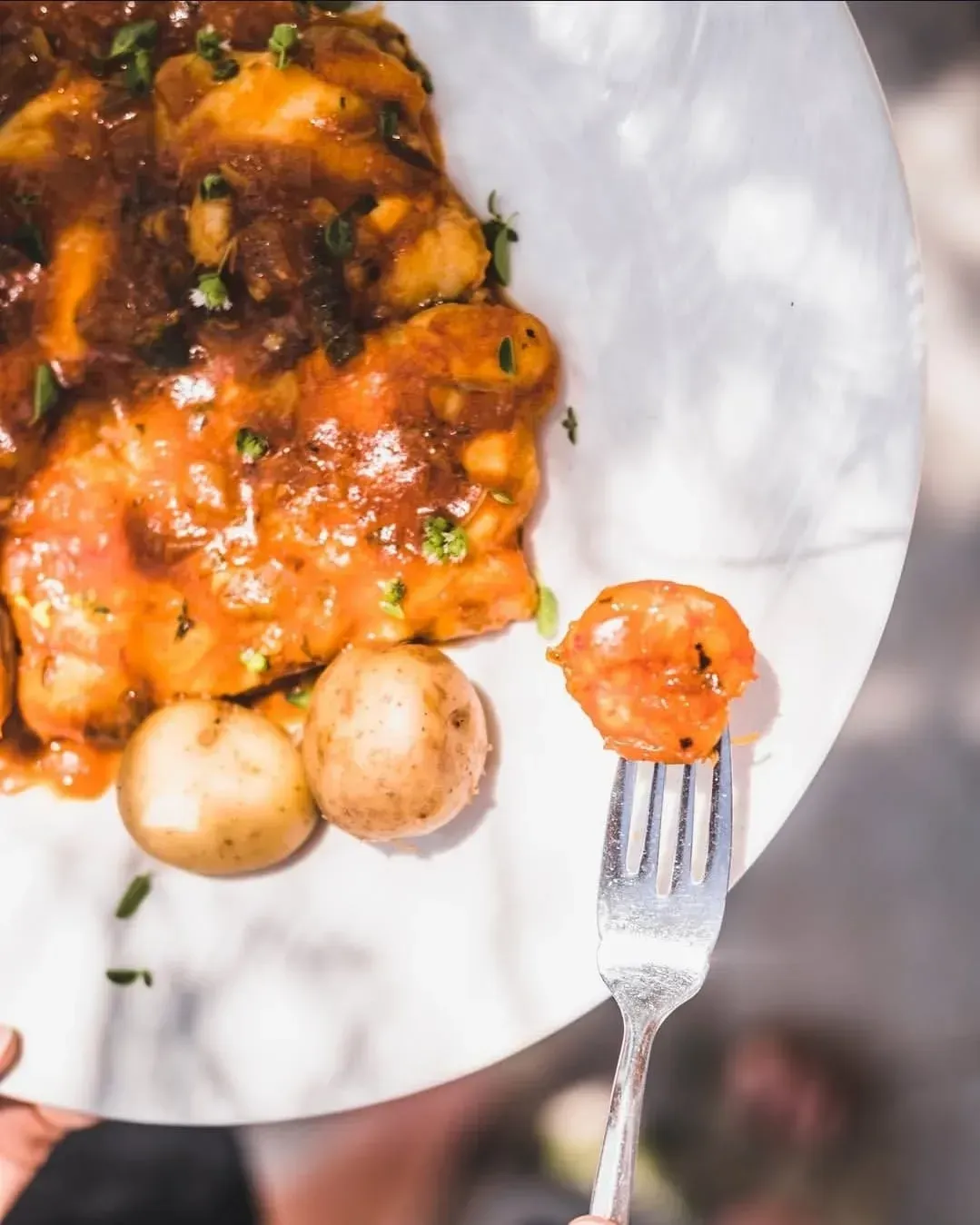 Un tenedor sostiene un solo camarón cocido sobre un plato blanco que contiene camarones en salsa roja y dos papas pequeñas hervidas.