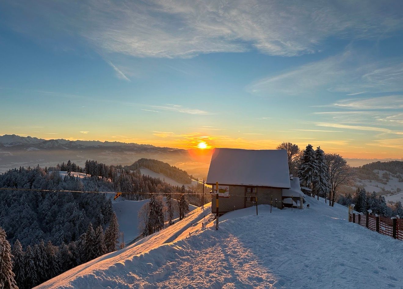Verschneite Berghütte im Sonnenaufgang unter leuchtend orangefarbenem Himmel und frostigen Bäumen