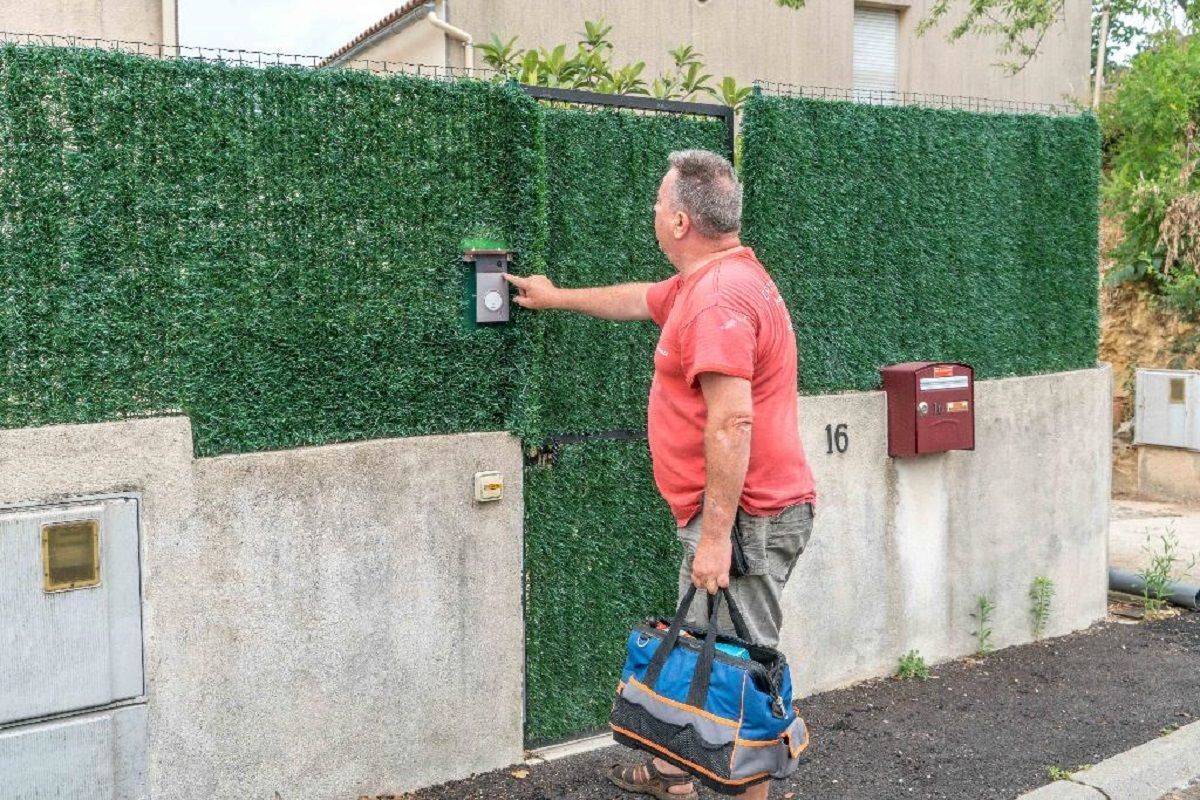 Une personne vêtue d'une chemise rouge et tenant une sacoche à outils appuie sur le bouton d'un interphone situé sur un portail recouvert d'une haie artificielle verte.