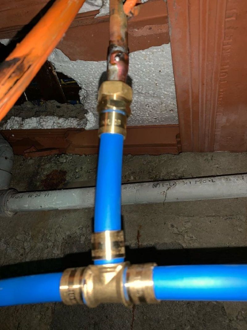 Un raccord de plomberie en forme de T reliant un tube PEX bleu à un tuyau de cuivre vertical avec des raccords en laiton dans un vide sanitaire sous plafond.