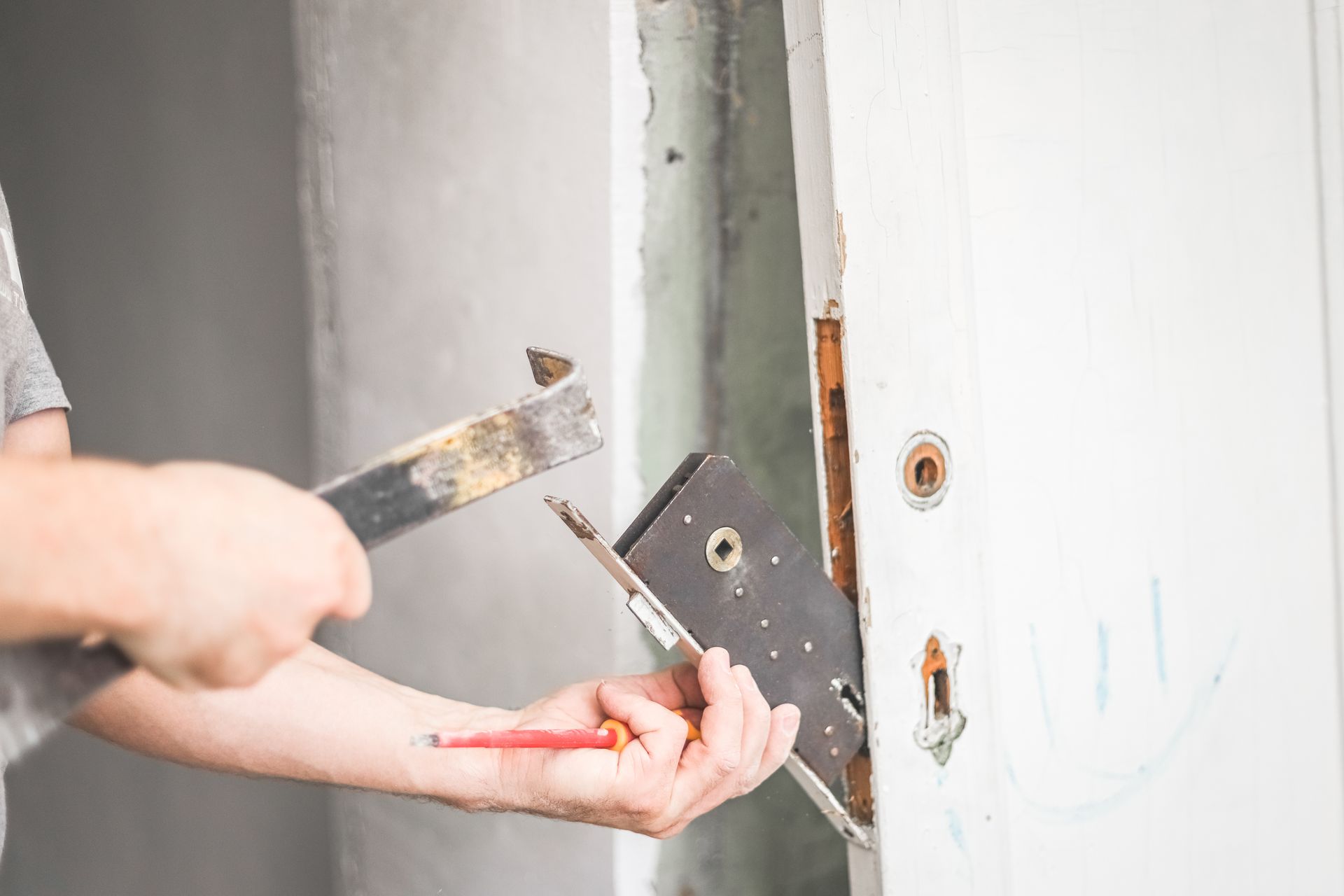 Une personne retire le mécanisme d'une serrure à mortaise du bord d'une porte blanche à l'aide d'un outil métallique et d'un tournevis.