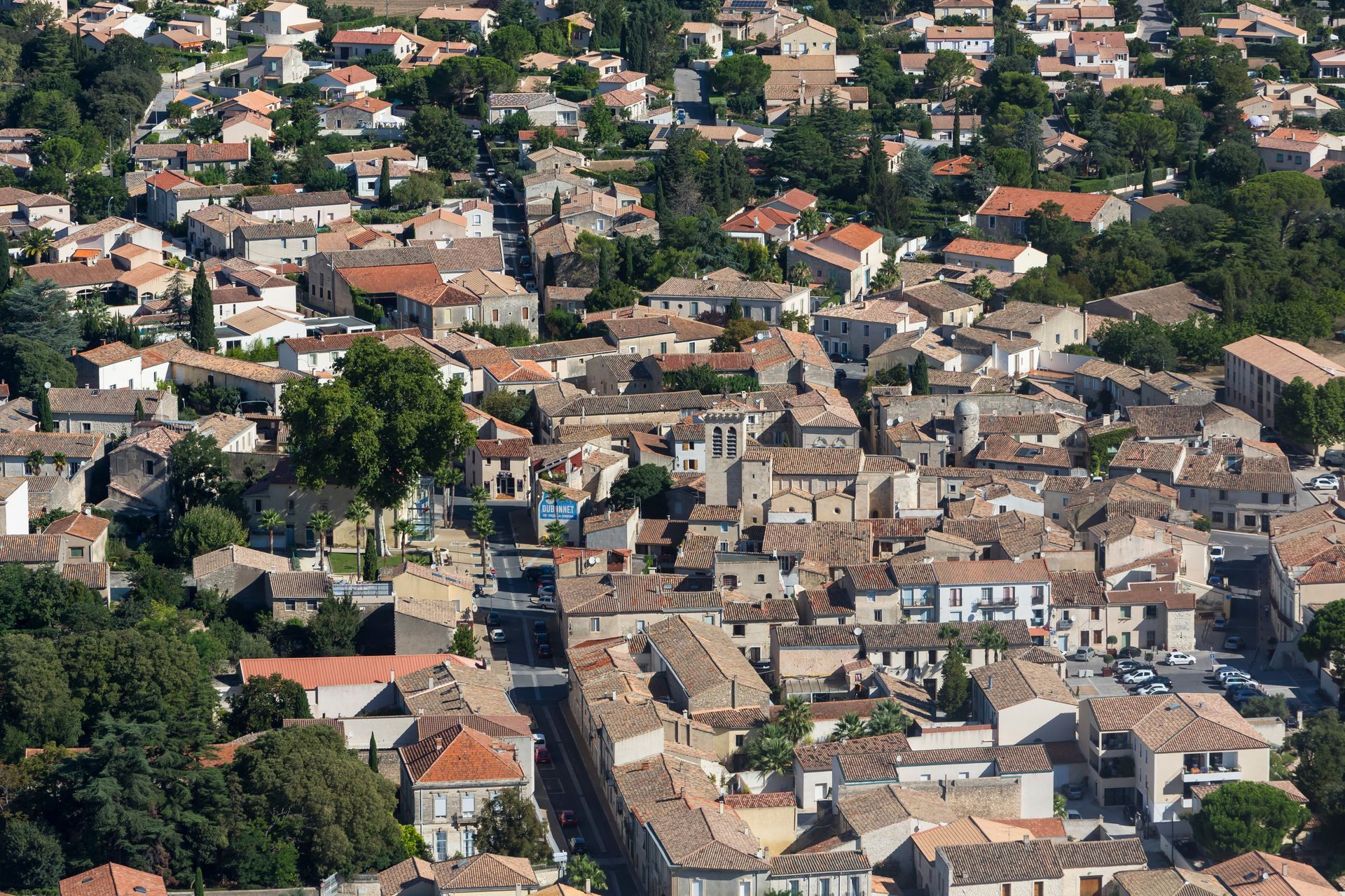 Vue aérienne d'une ville méditerranéenne dense et ensoleillée, avec des toits de tuiles, des bâtiments en pierre et des arbres verdoyants.