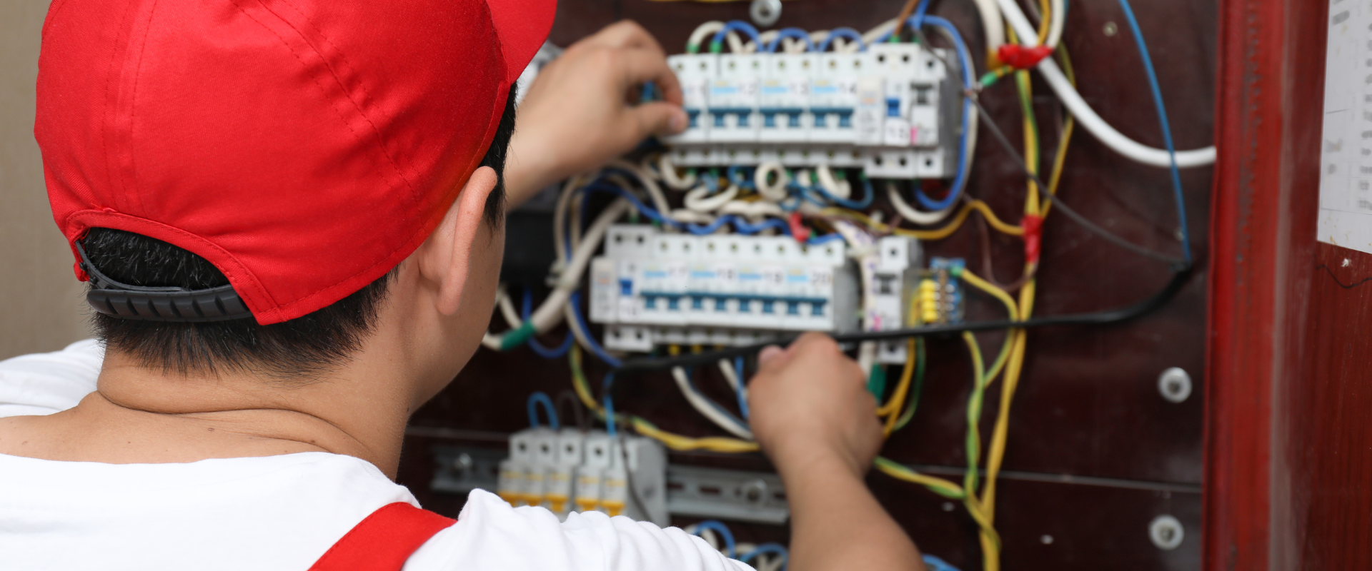 Électricien travaillant sur un tableau électrique, portant une casquette rouge et une chemise blanche.