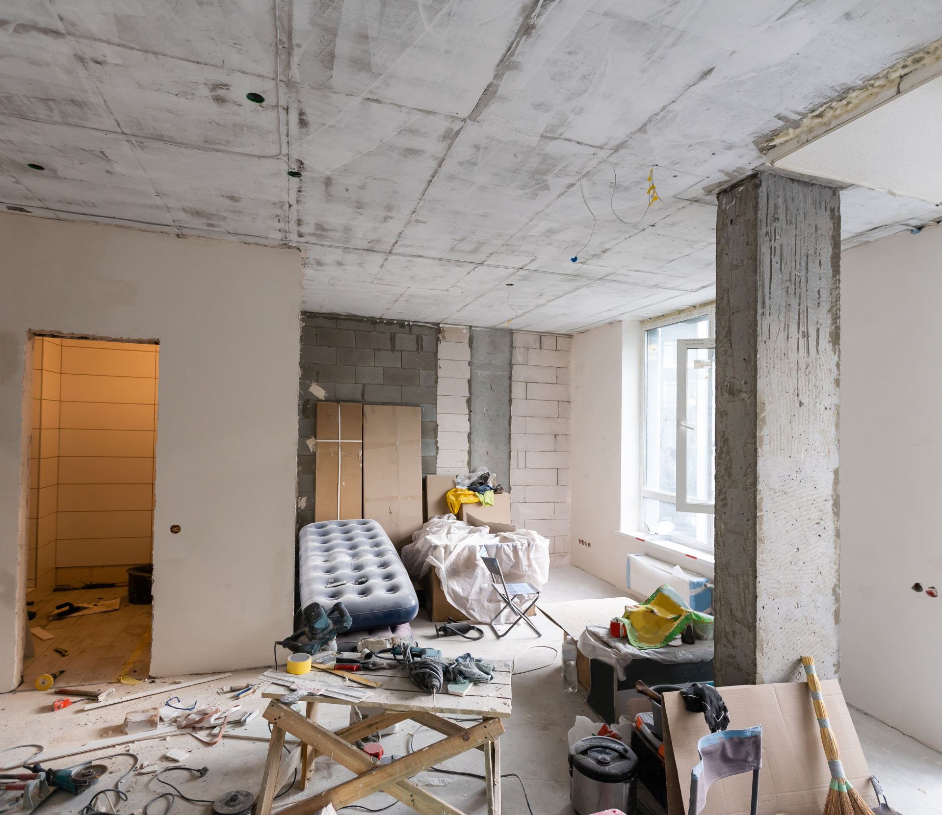 Intérieur d'une pièce en construction, avec du béton apparent, des outils et des débris éparpillés.