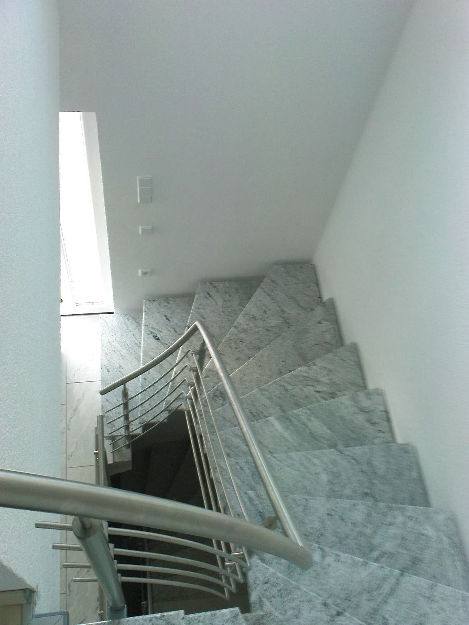 Treppe mit grauen Steinstufen, Metallgeländer, weissen Wänden und heller Deckenbeleuchtung.
