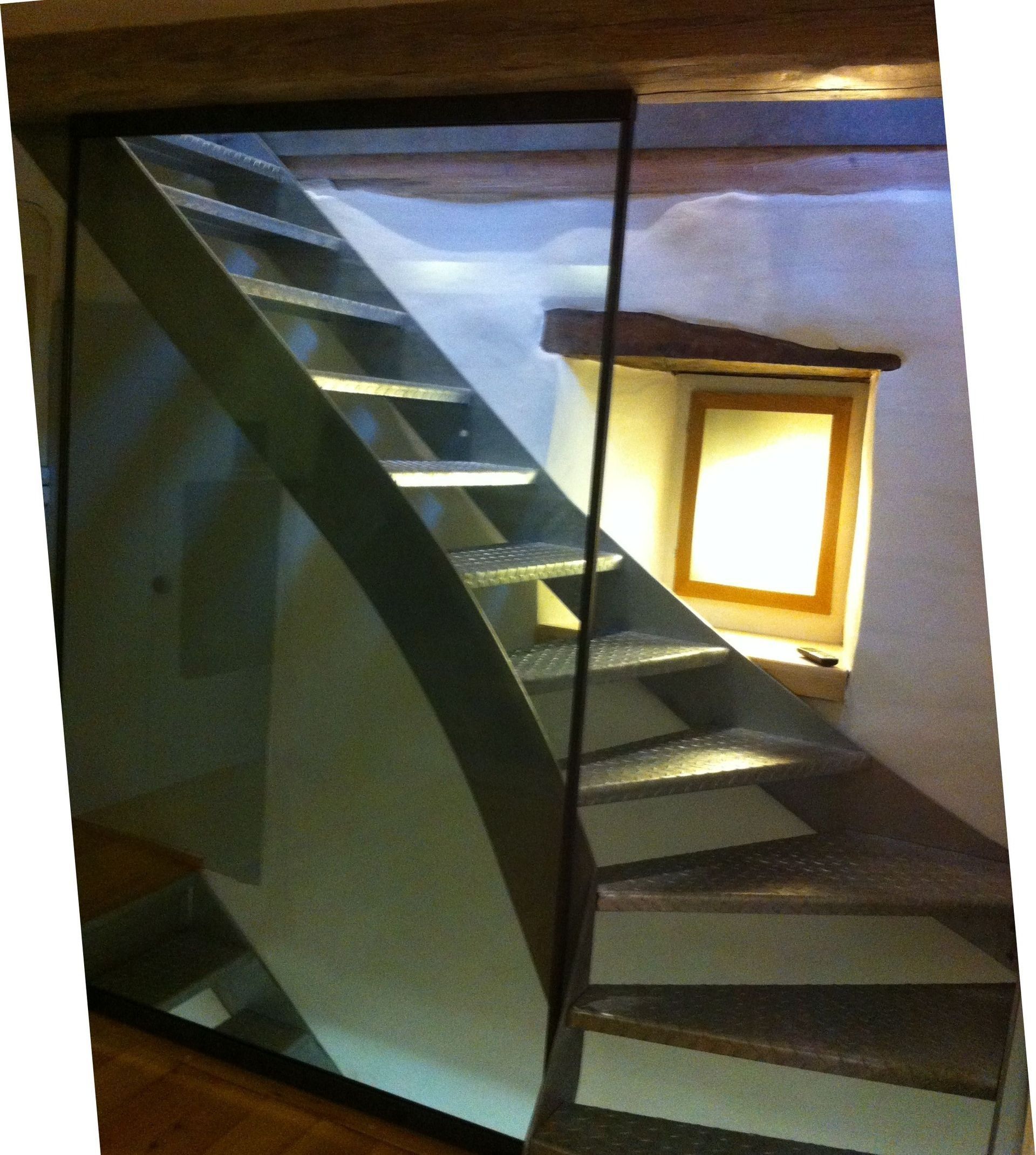 Eine Metalltreppe mit seitlicher Glaswand vor einer hellen Wand führt zu einem Fenster hinauf.