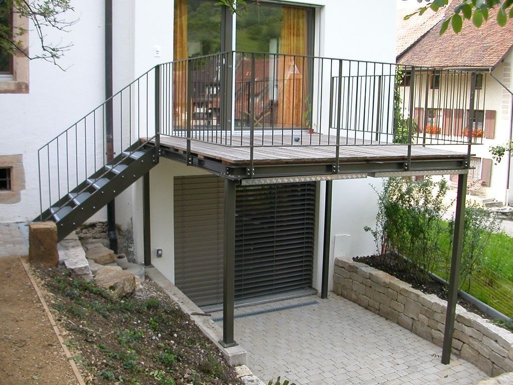 Metallbalkon mit Treppe über einem ebenerdigen Garagentor. Eine Steinmauer umgibt einen kleinen Garten.