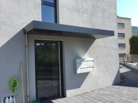 Dunkelgraue Markise über der Haustür, weisser Briefkasten, graues Gebäude, sonniger Tag.