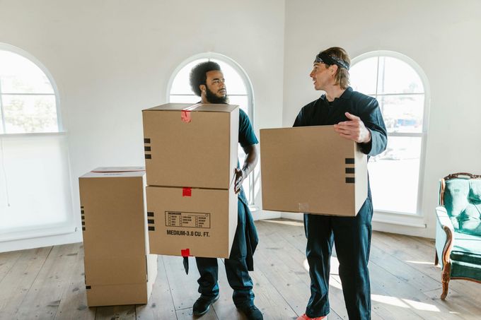 Dos personas con uniformes de trabajo oscuros transportan cajas de cartón para mudanzas en una habitación luminosa y escasamente amueblada, con ventanas arqueadas.