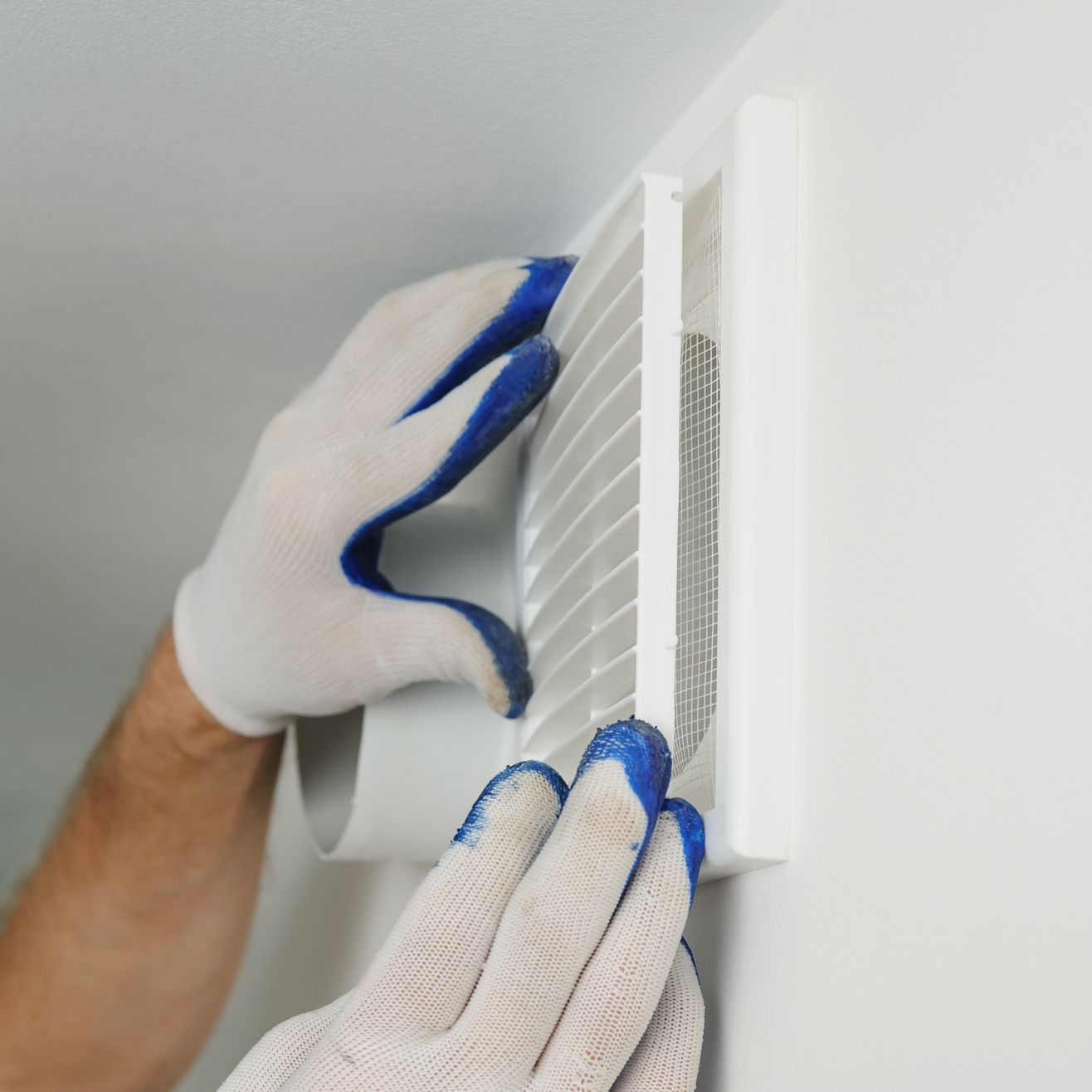 Des mains gantées fixent une grille d'aération blanche au mur.