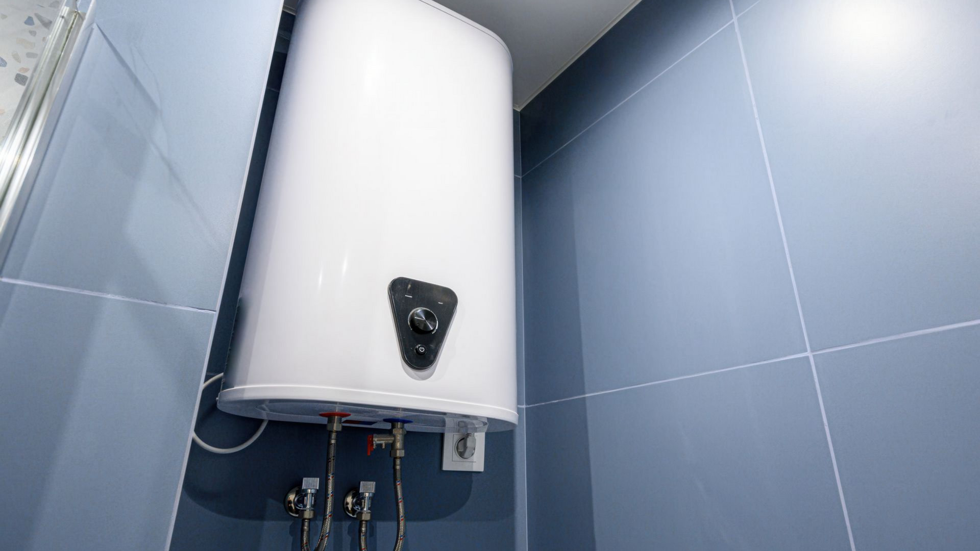 Chauffe-eau blanc sur un mur carrelé bleu, laissant apparaître les tuyaux et le panneau de commande.