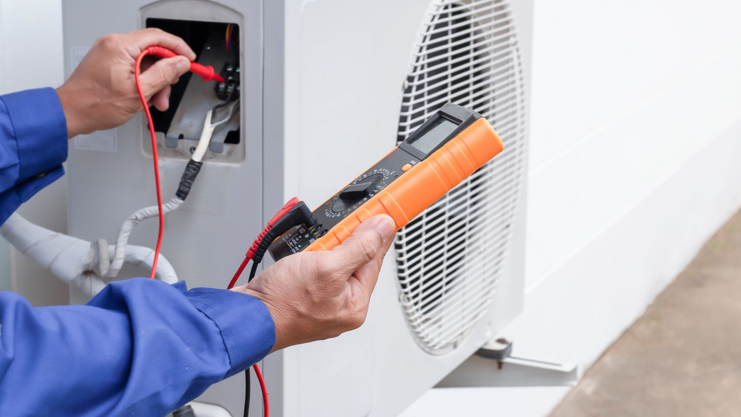 Une personne en salopette bleue utilise un multimètre pour inspecter une unité de climatisation extérieure.