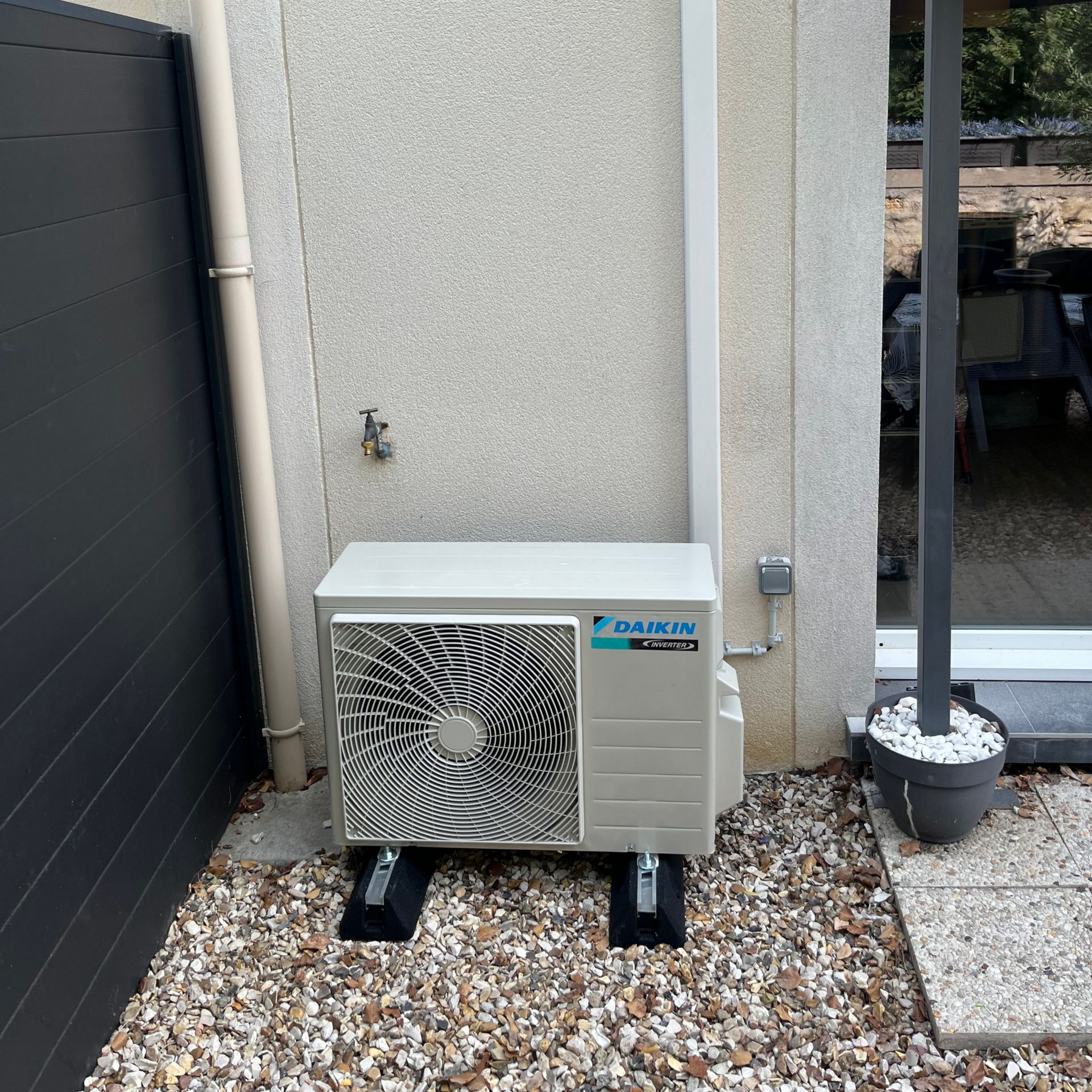 Un climatiseur posé sur un lit de gravier, monté sur des blocs noirs, à côté d'un mur.