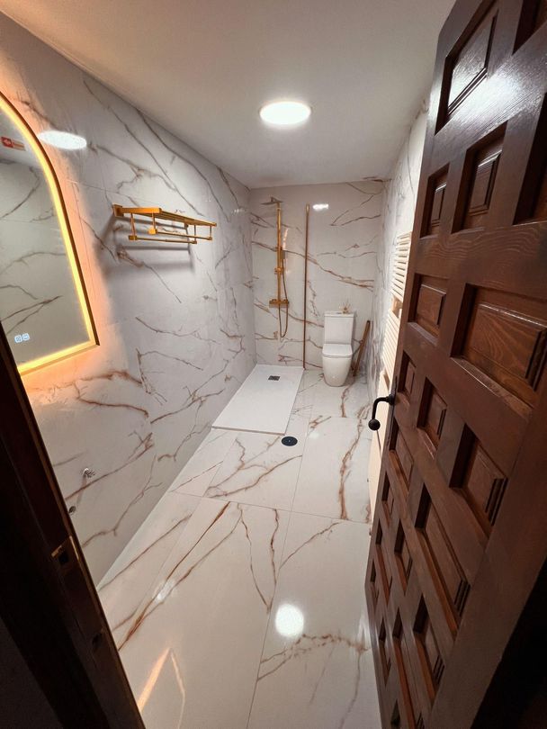 Baño moderno de mármol con inodoro, espejo de pared, grifería de ducha y tocador iluminado.