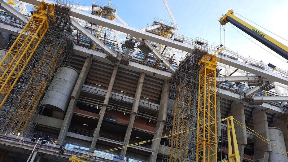 Gran obra de construcción industrial con estructura de acero, grúas y andamios amarillos bajo un cielo nublado.