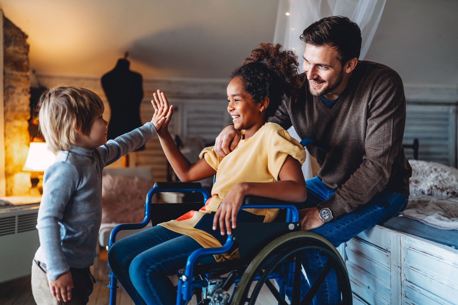 Une fille en fauteuil roulant fait un high-five à un garçon tandis qu'un homme sourit, dans une chambre.