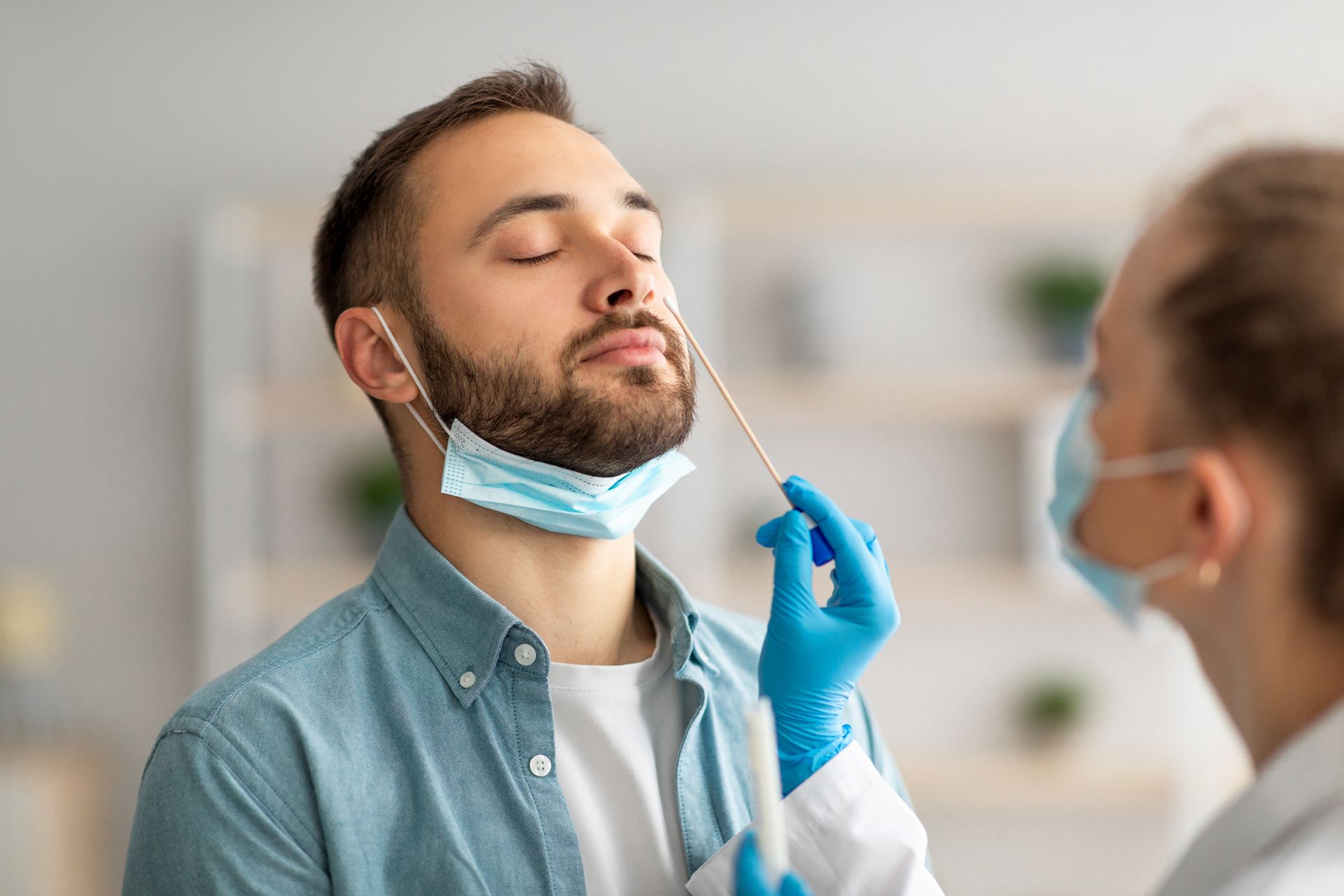 Un homme se soumet à un test par écouvillonnage nasal à l'intérieur, les yeux fermés, masque sous le menton. Un soignant porte des gants et un masque.