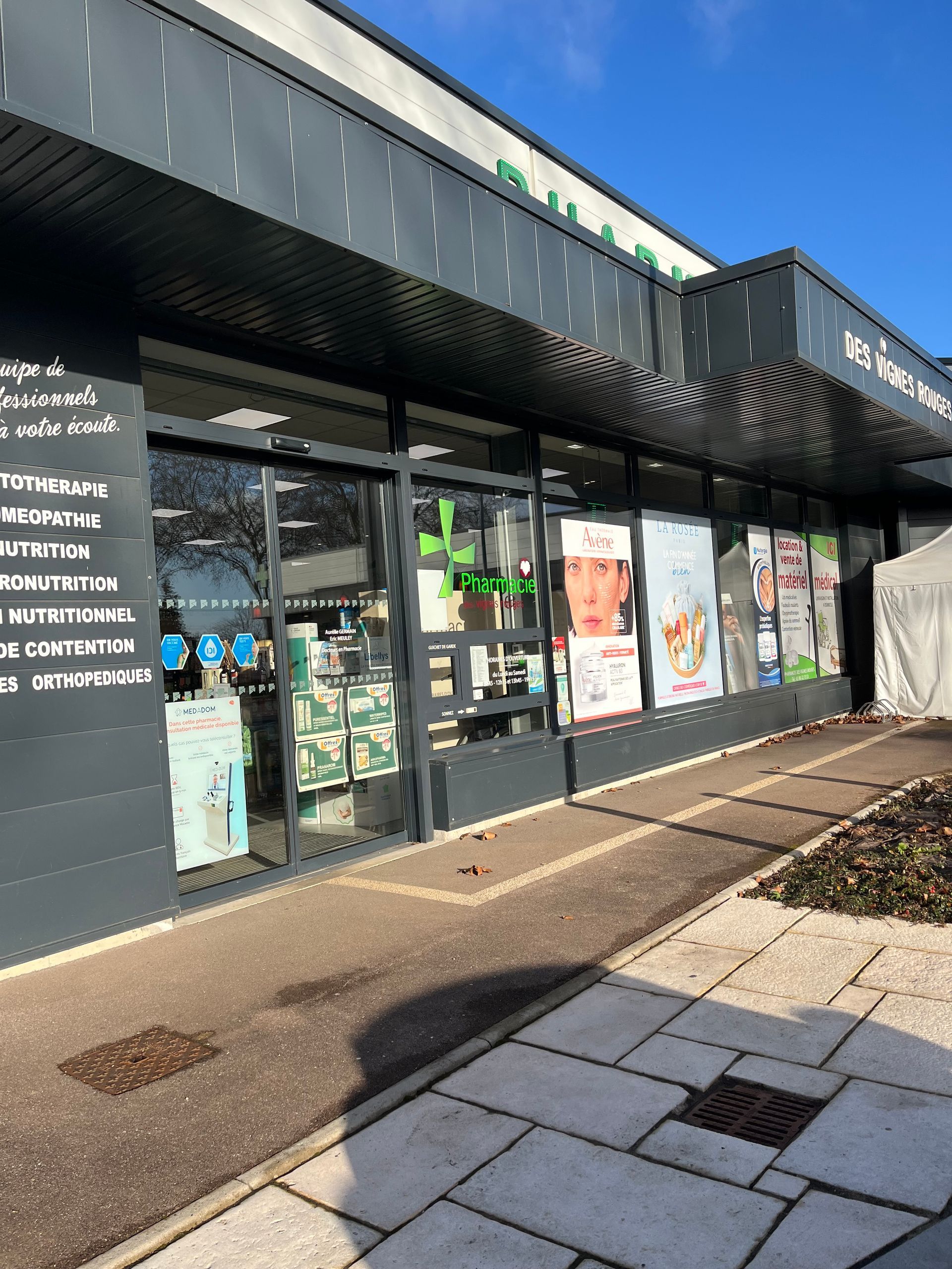 Devanture de pharmacie avec une façade gris foncé, des vitrines et des présentoirs de produits.