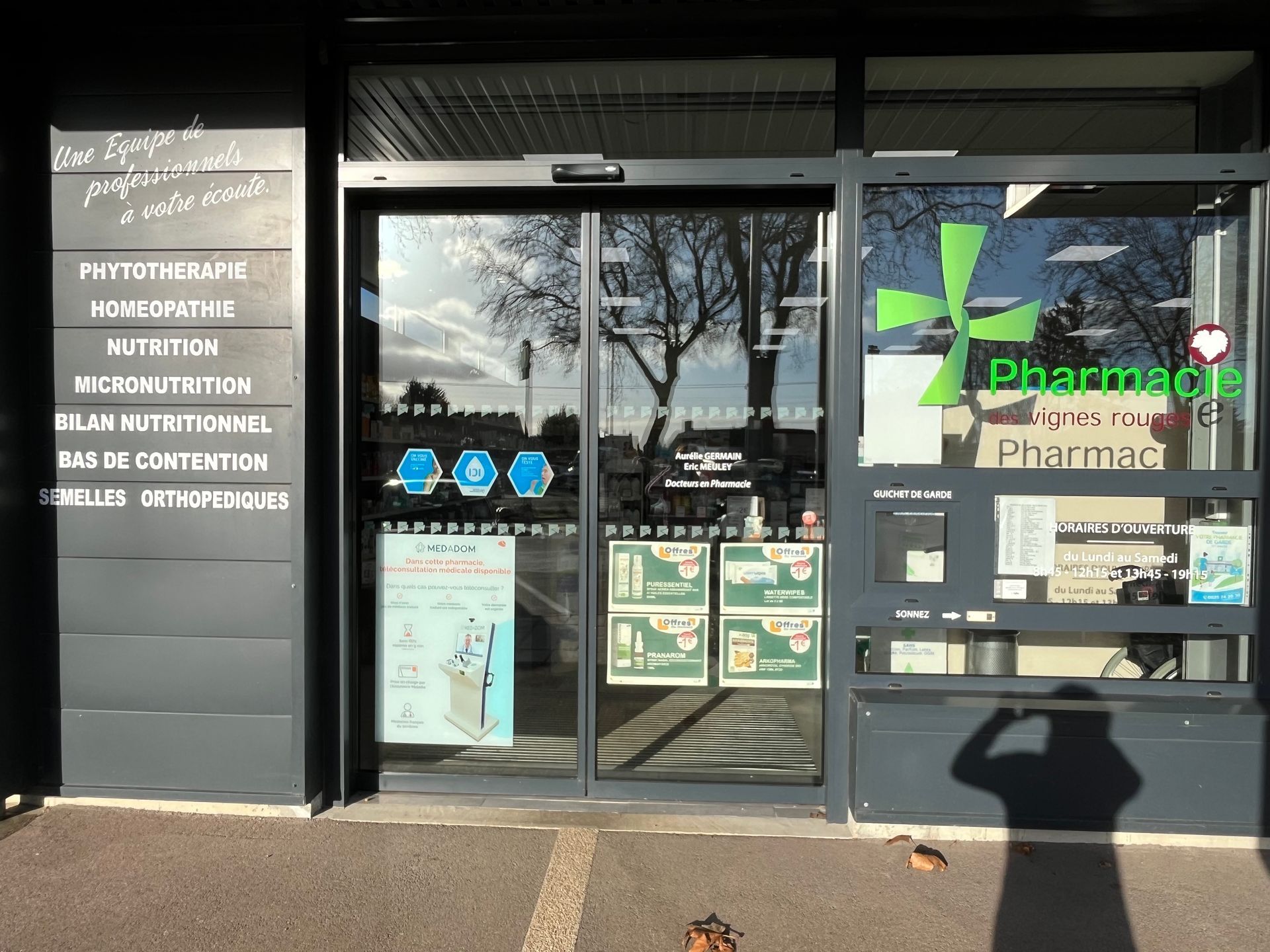 Entrée de la pharmacie avec portes coulissantes automatiques; logo croix verte, divers affichages.