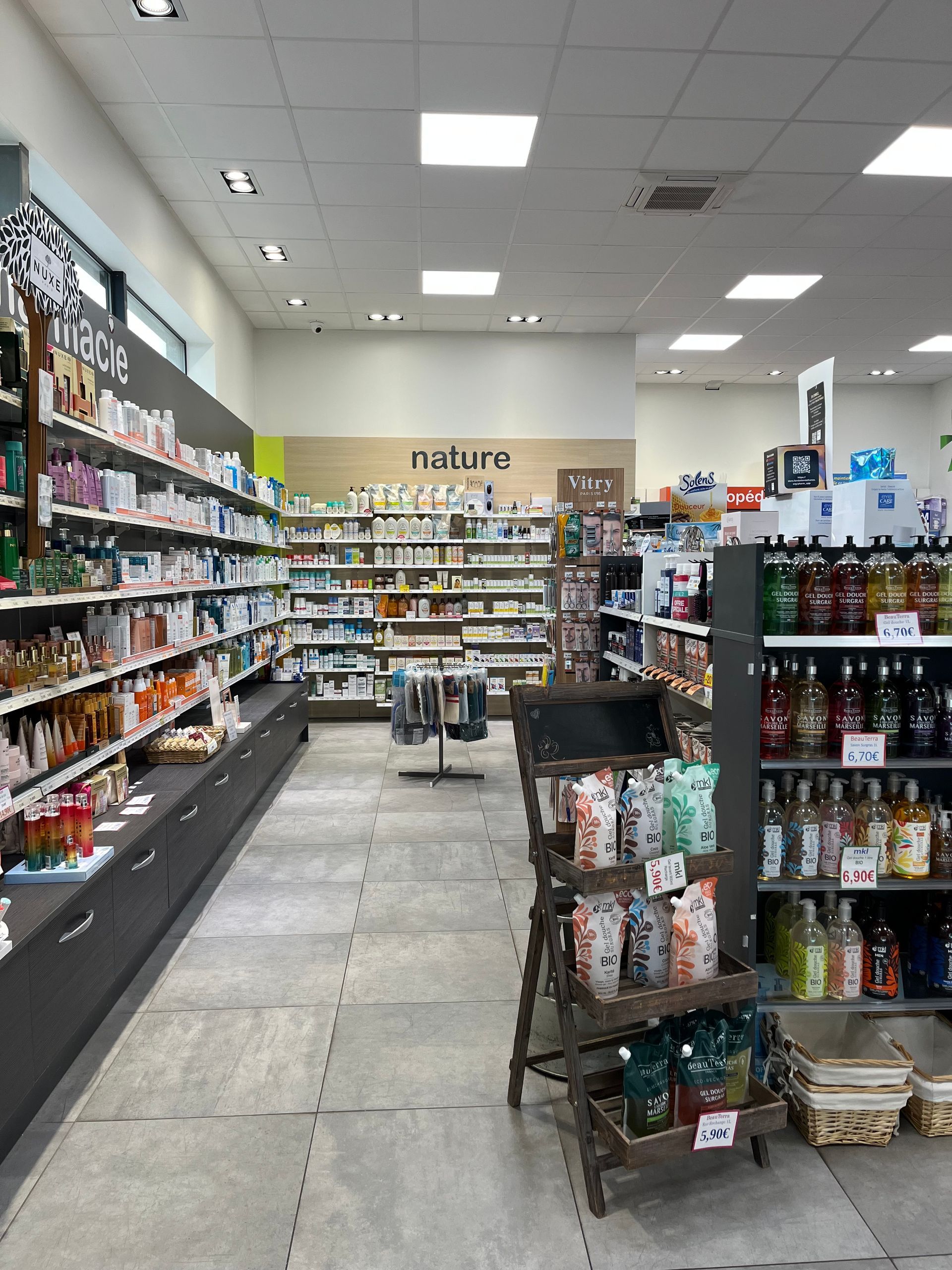 Intérieur d'un magasin de produits de santé ; étagères remplies de produits, panneau « nature » sur le mur du fond, allée avec présentoir.