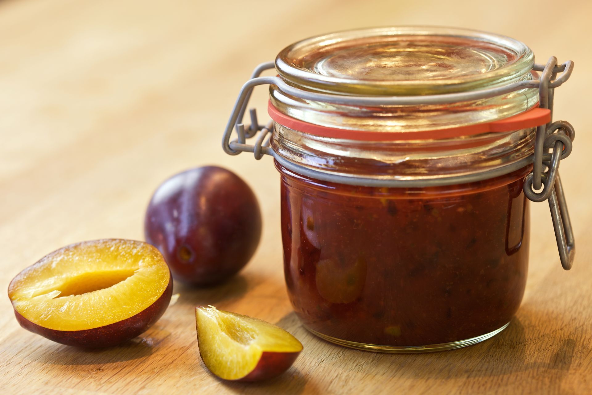 A jar of plum jam sits on a wooden table next to plums
