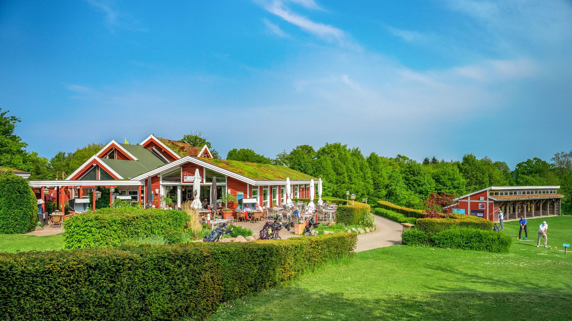 Ein rotes Haus mit grünem Dach ist von Bäumen und einer Wiese umgeben.