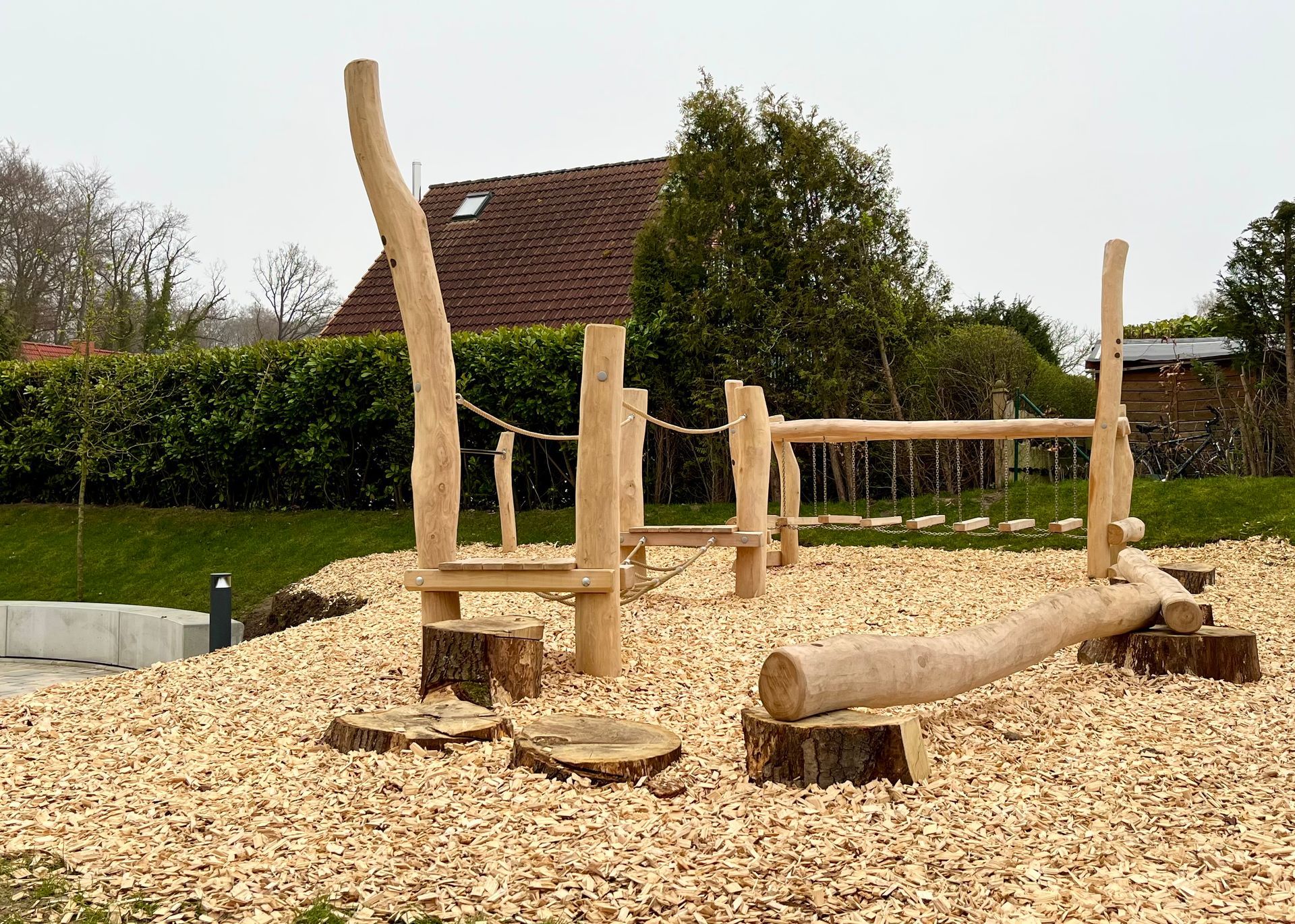 Spielplatz aus Holz mit Stufen aus Baumstämmen, Balancierbalken und Klettergerüst auf einem Holzschnitzelbelag.