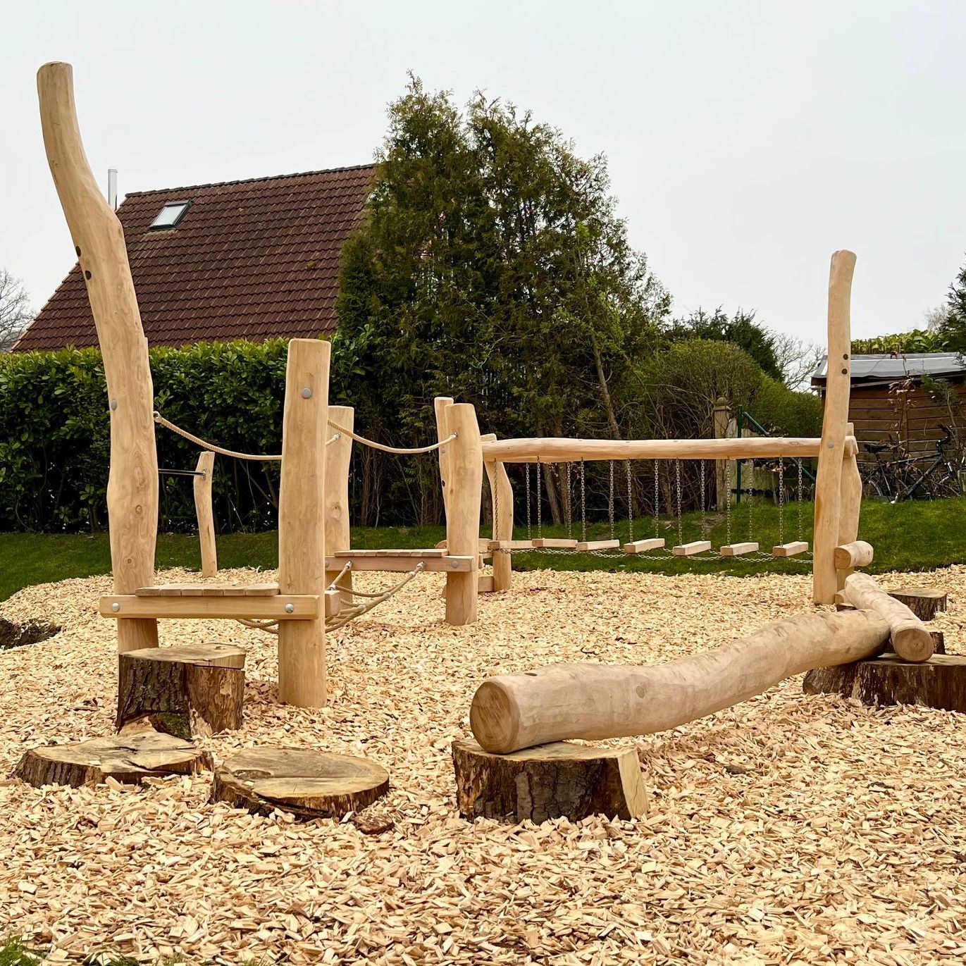 A wooden playground is surrounded by wood chips and a house in the background.